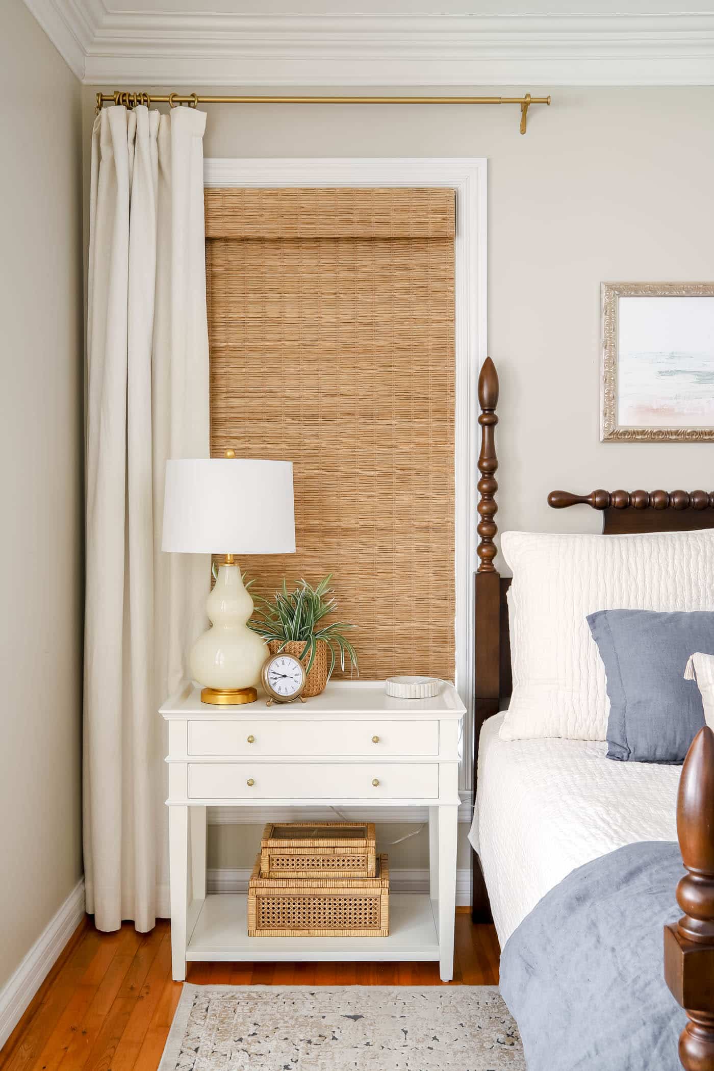 nightstand with lamp, clock and decorative boxes with ivory curtains, bamboo shades and revere pewter was in bedroom.