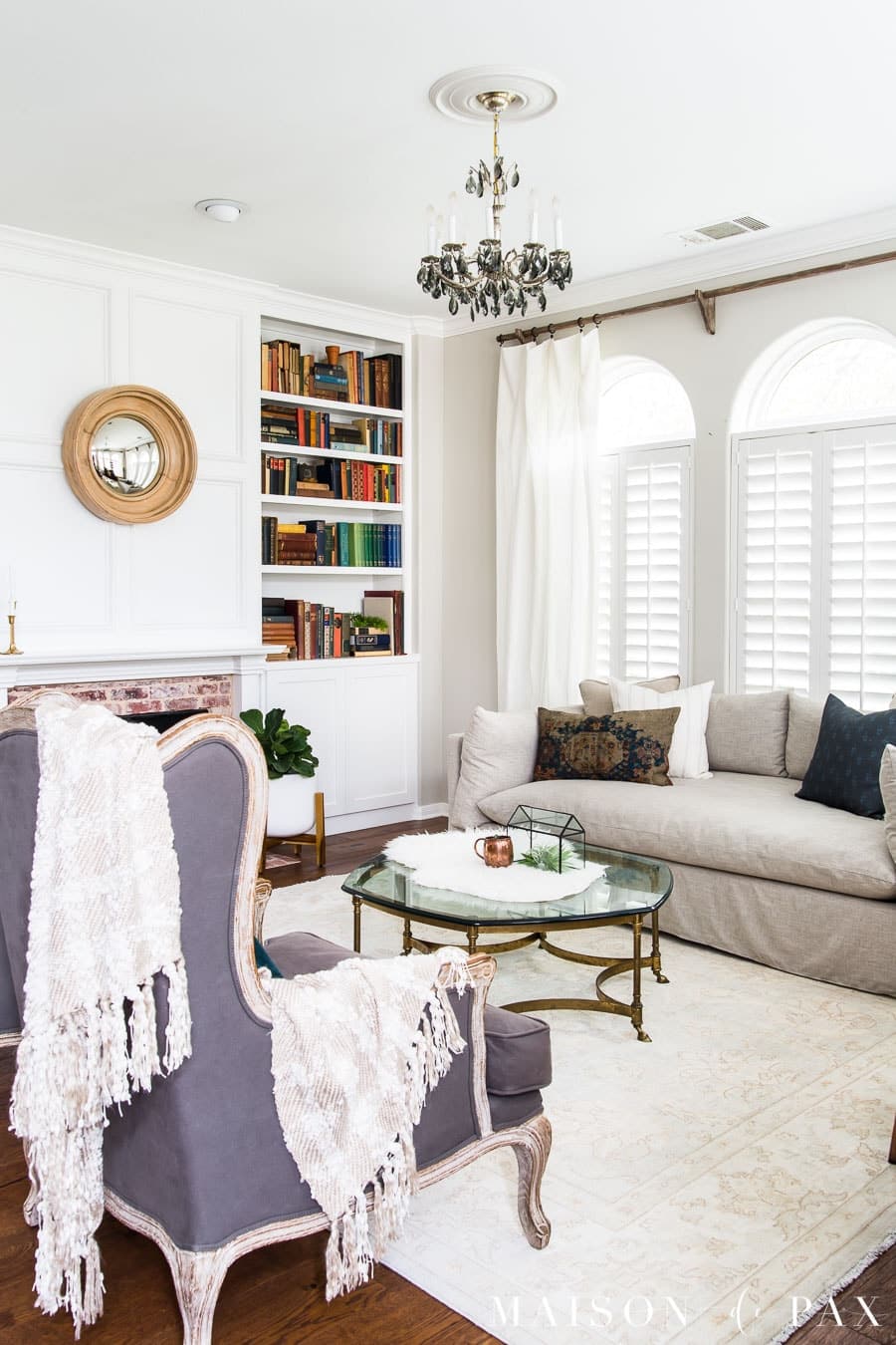 Neutral transitional living room with a slipcovered sofa, built-in bookshelves, white fireplace, glass coffee table, and arched windows with plantation shutters.