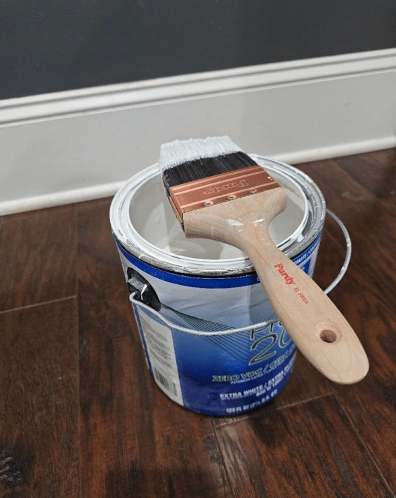Paintbrush resting on an open can of white paint on a hardwood floor before painting a room.
